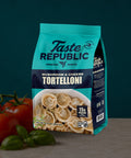 A bag of Taste Republic Mushroom & Cheese Tortelloni pasta stands on a countertop with tomatoes and basil leaves beside it. The package is teal and dark blue with a photo of the cooked tortelloni in a bowl on the front.